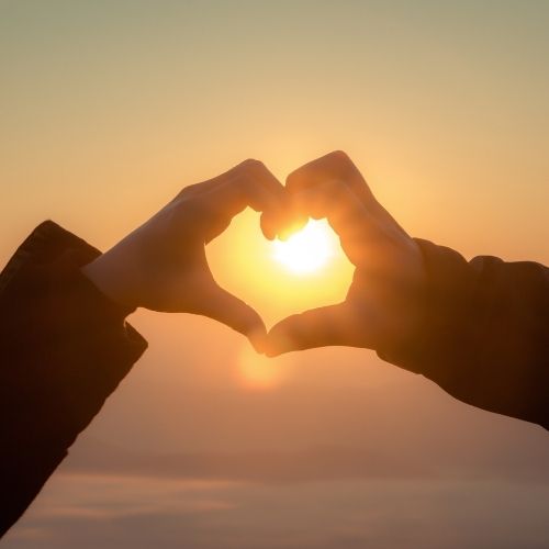 Two hands form a heart shape against a bright sunlit background, symbolizing love and warmth