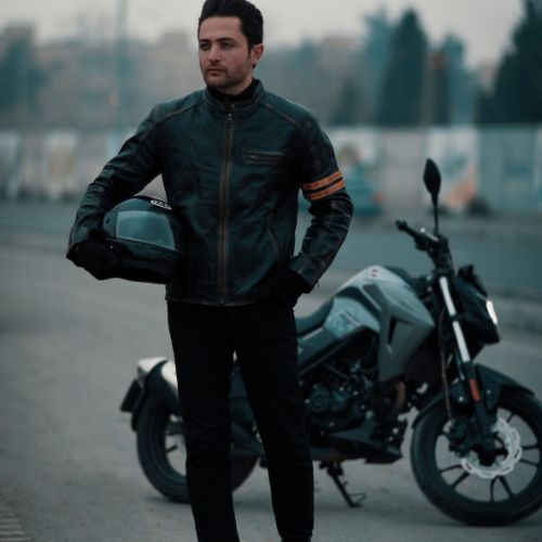 A man in a black leather jacket stands beside a motorcycle