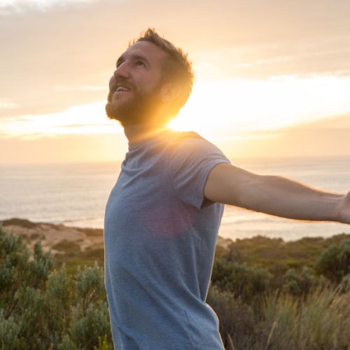 A man stands on a hill at sunset, arms outstretched