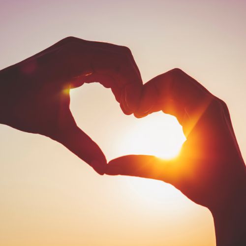 Two hands form a heart shape against a bright sunlit background